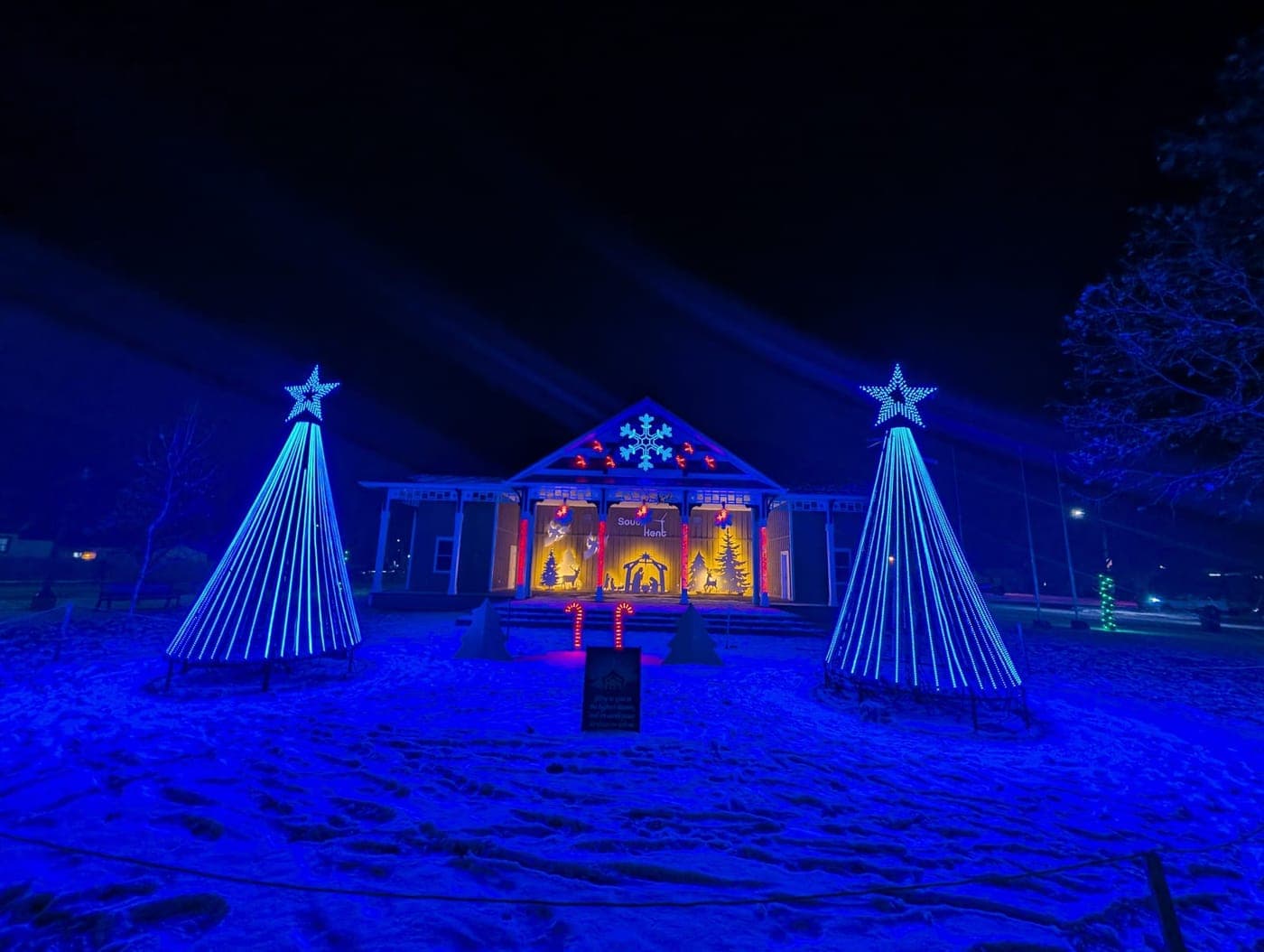 Outdoor Christmas display with two blue light-cone trees flanking a warmly lit nativity scene at a community pavilion.