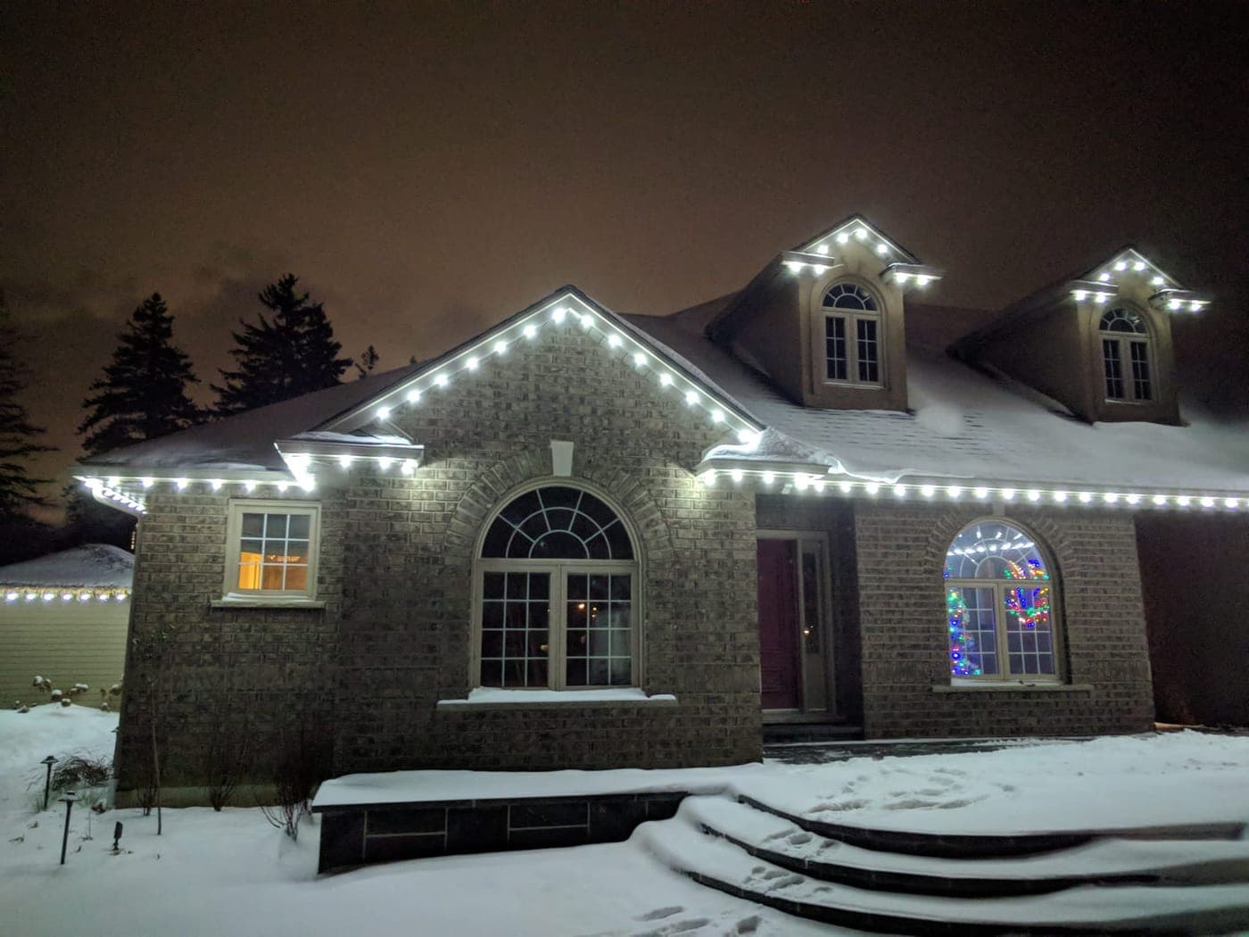 Front of a brick home lined with white roofline Christmas lights on a snowy winter night