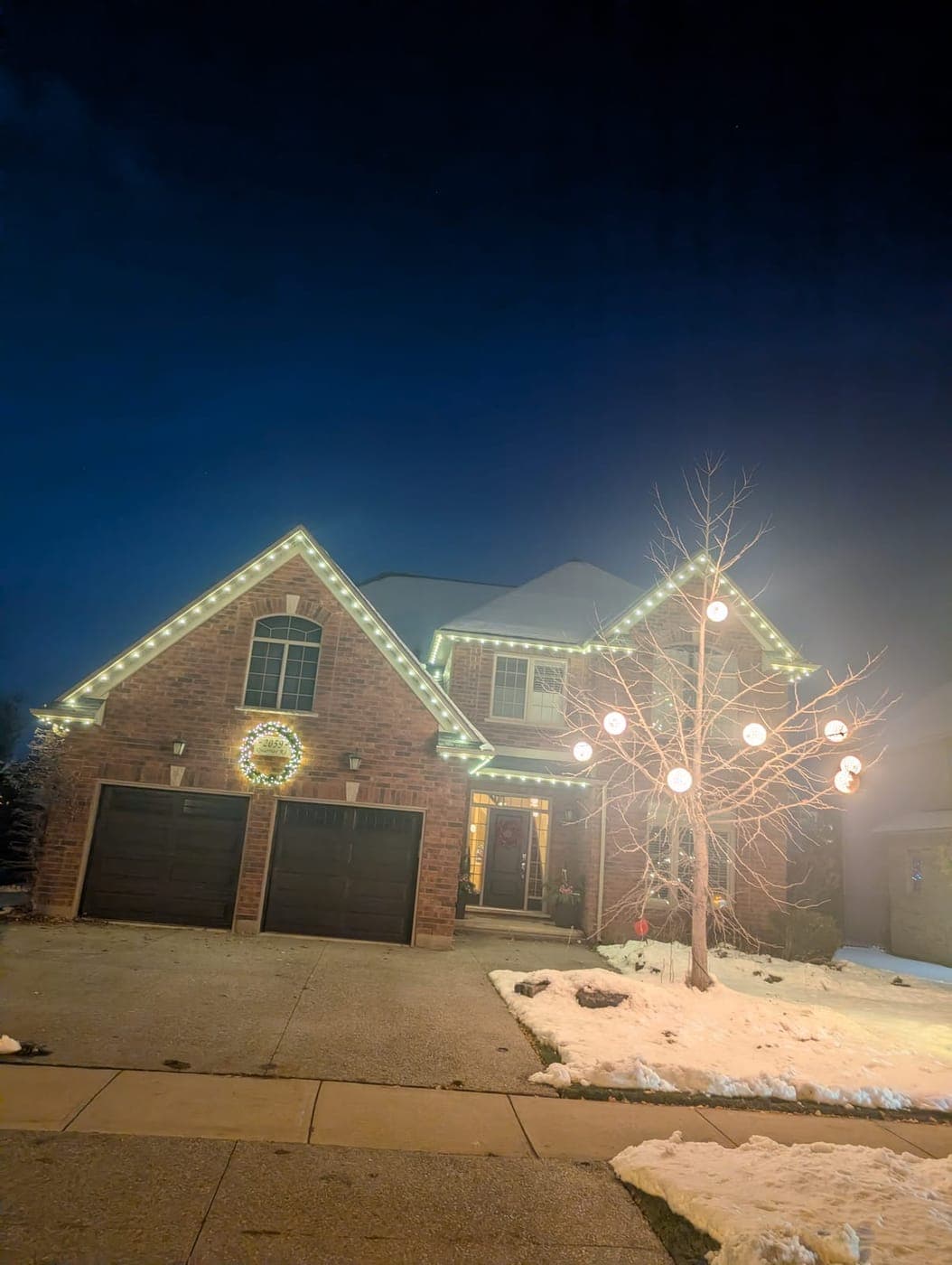 Brick home with warm-white roofline lights, a garage wreath and an illuminated globe-lit tree at night