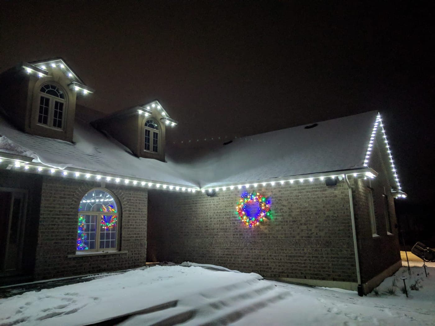 Brick home with white roofline Christmas lights across the dormers and a lit wreath in winter snow