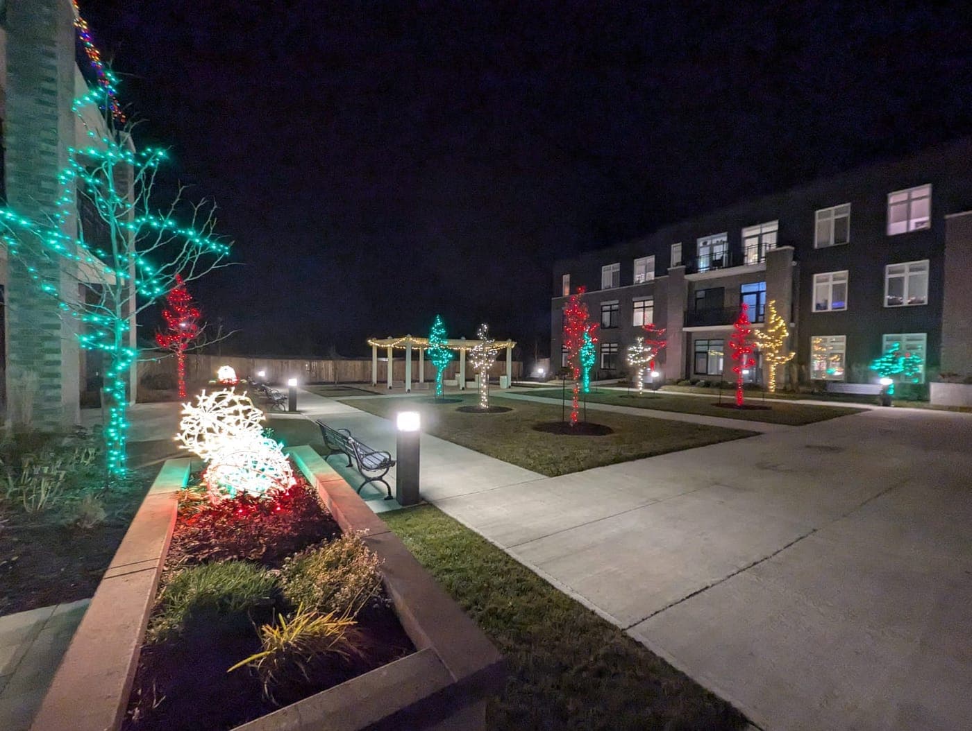 Commercial courtyard walkway with red and green light-wrapped trees and a lit holiday ornament at night