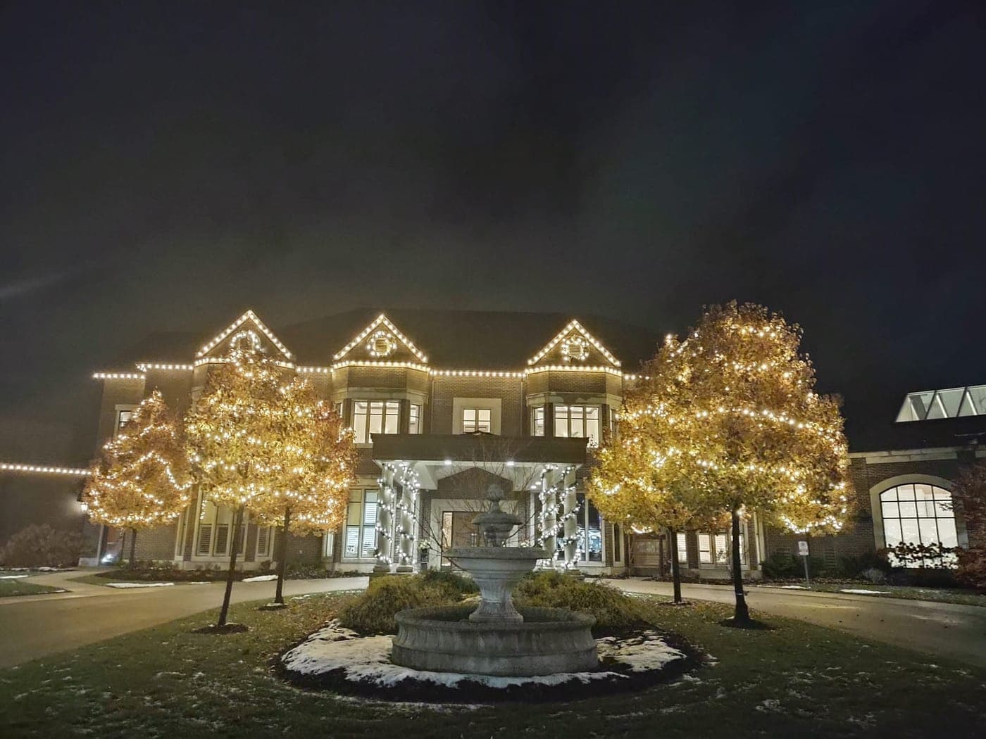 Grand event venue with warm-white roofline Christmas lights and tree wrapping around a stone fountain