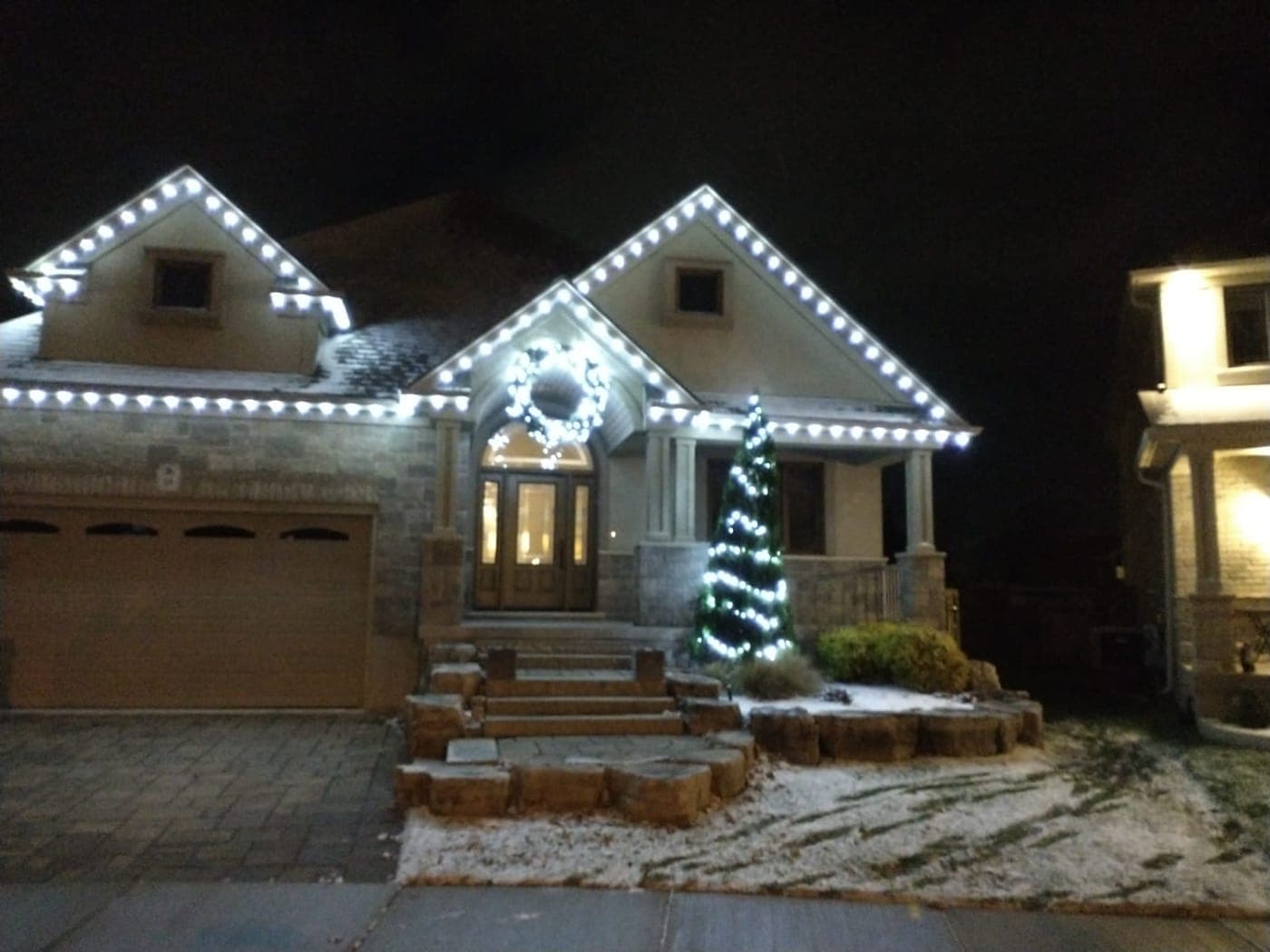 Cool-white C9 roofline lights and a lit wreath on a stone bungalow with a spiral cone tree in the snow.
