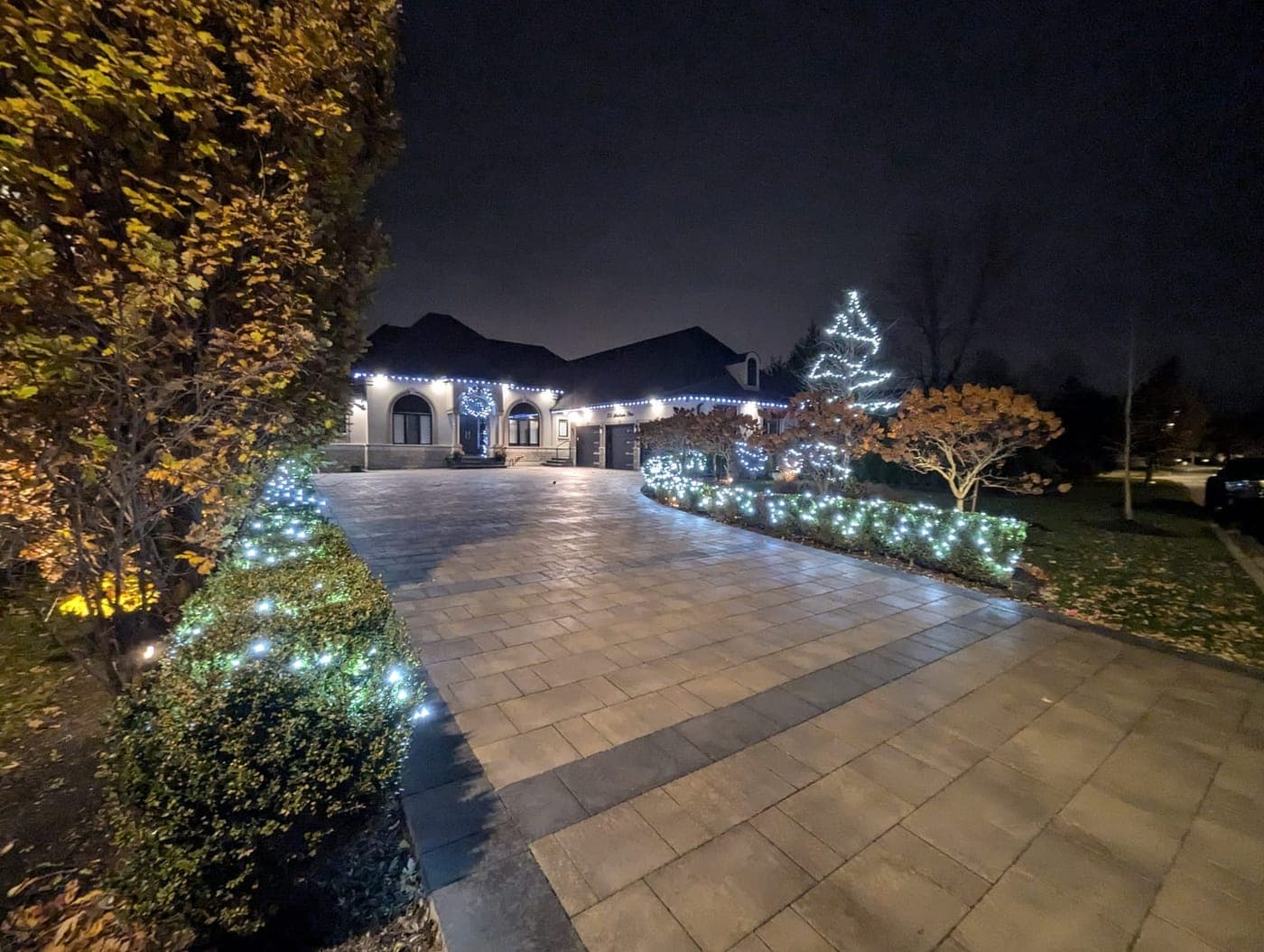 Estate driveway lined with cool-white lit shrubs leading to a home with roofline Christmas lights