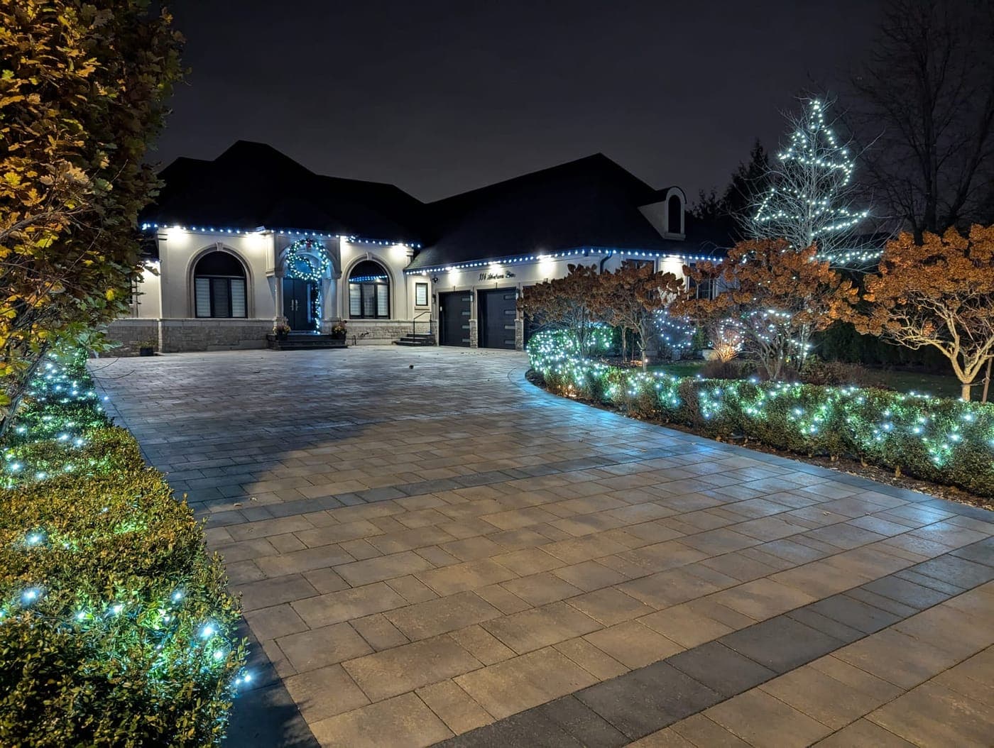 Estate home outlined in white Christmas lights with a light-wrapped tree and glowing shrub beds