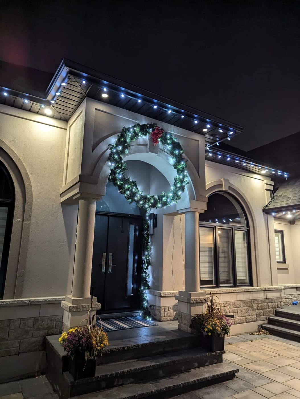 Lit Christmas wreath and garland draped over a stone portico entrance with blue soffit accent lights