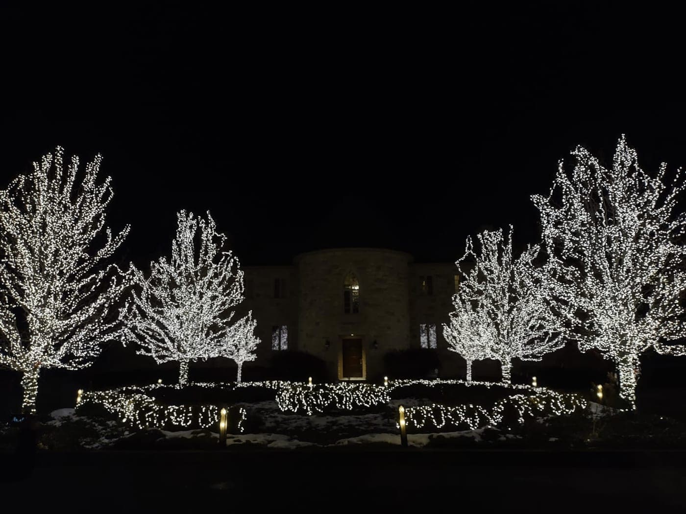 Symmetrical stone estate front with four large trees in dazzling cool-white Christmas lights