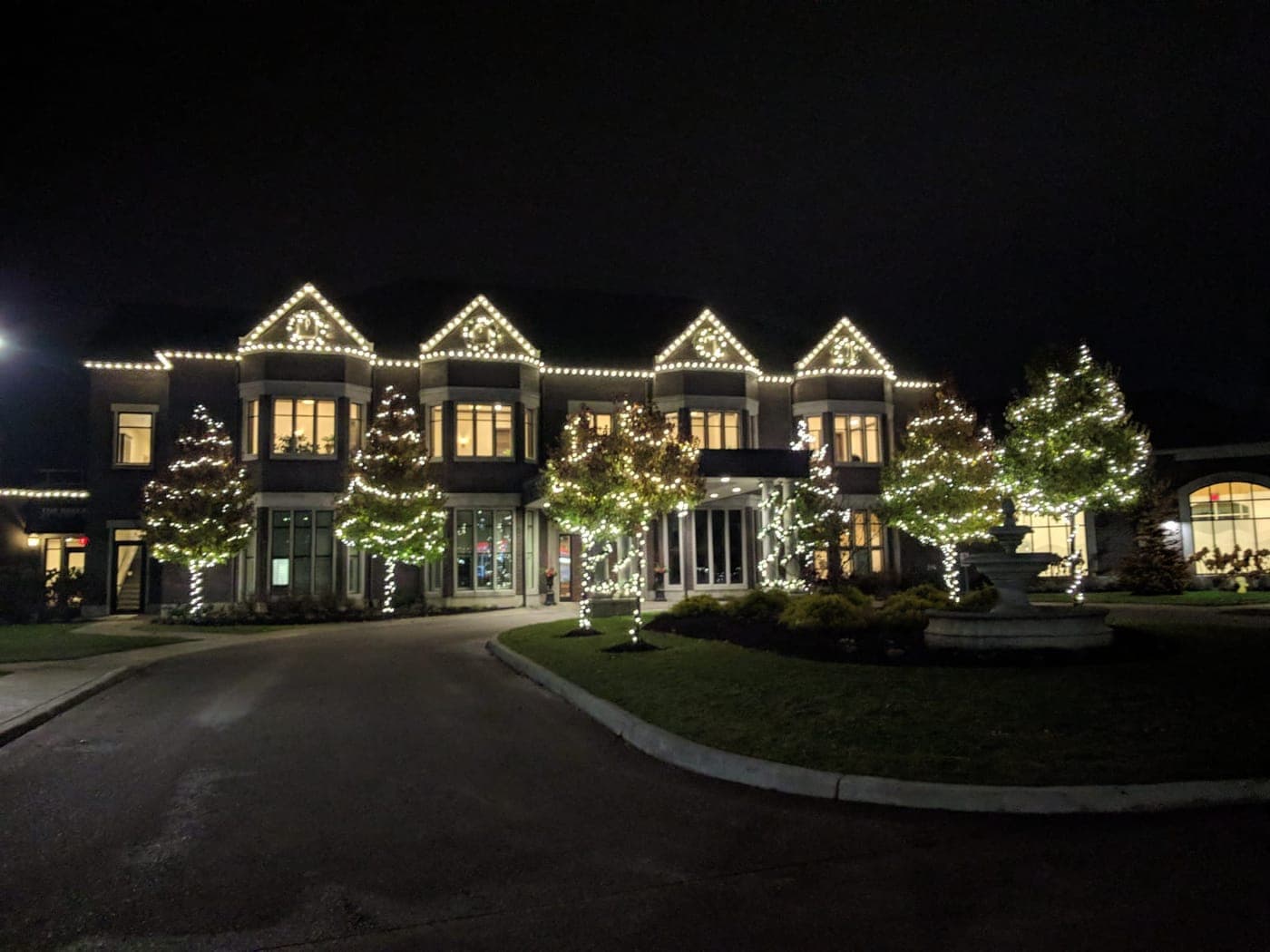 Large estate building illuminated with warm-white holiday roofline lighting and lit landscape trees.