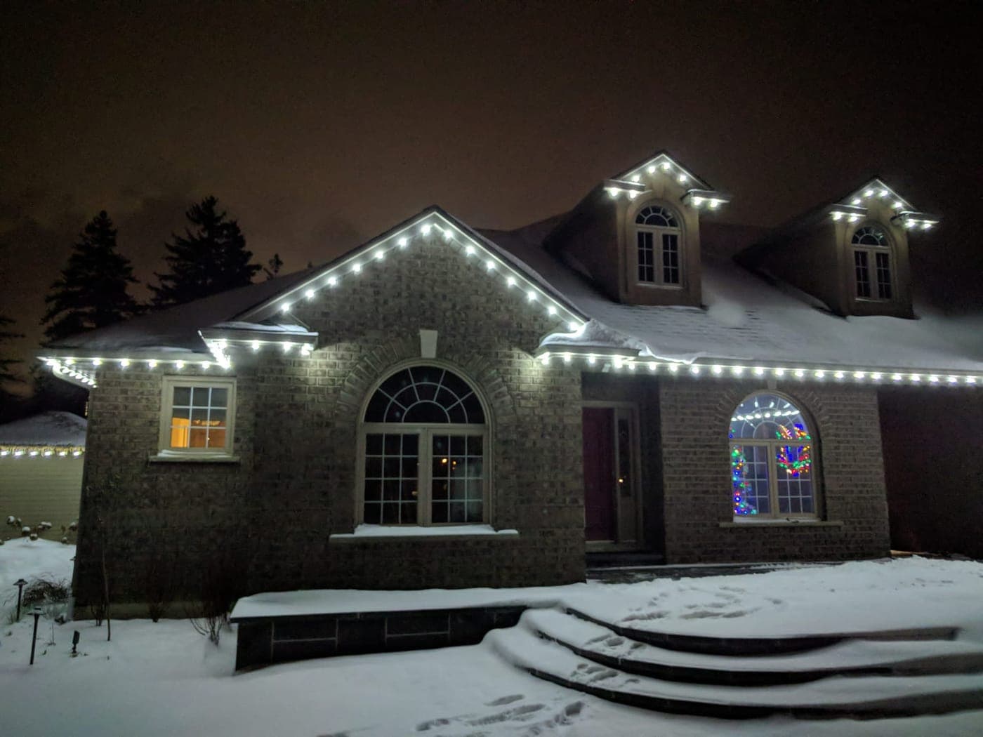 Grey brick home with white roofline lights and colourful lit wreaths glowing in the arched windows