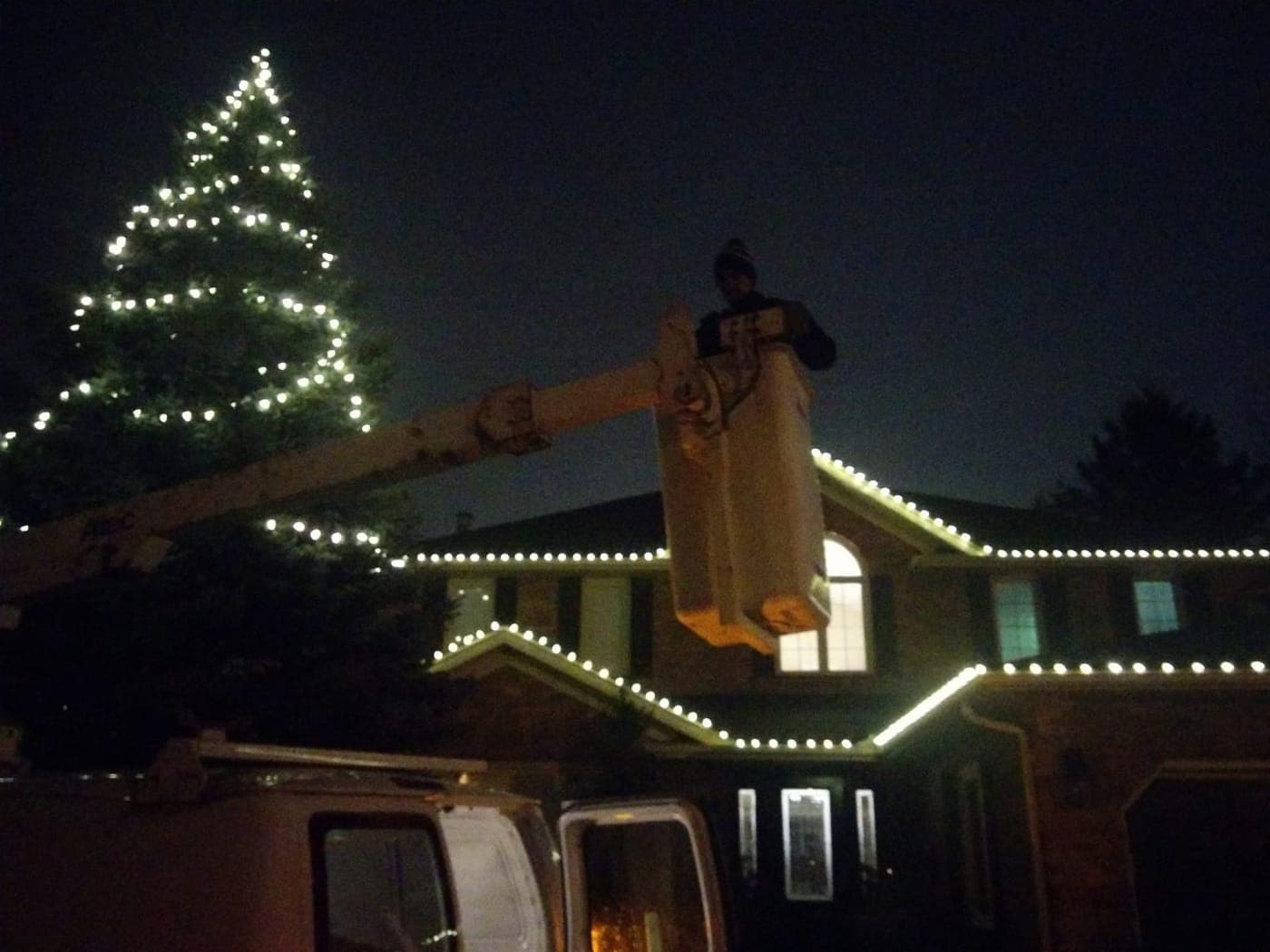 Christmas light installer working from an aerial bucket to hang warm white lights on a brick home's roofline.