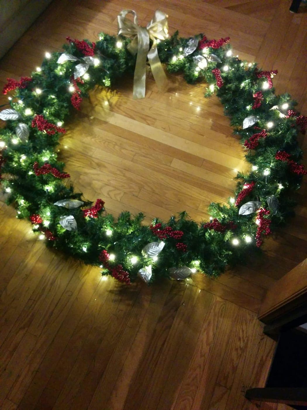 Pre-lit Christmas wreath with red berries, silver leaves and a gold bow on a hardwood floor