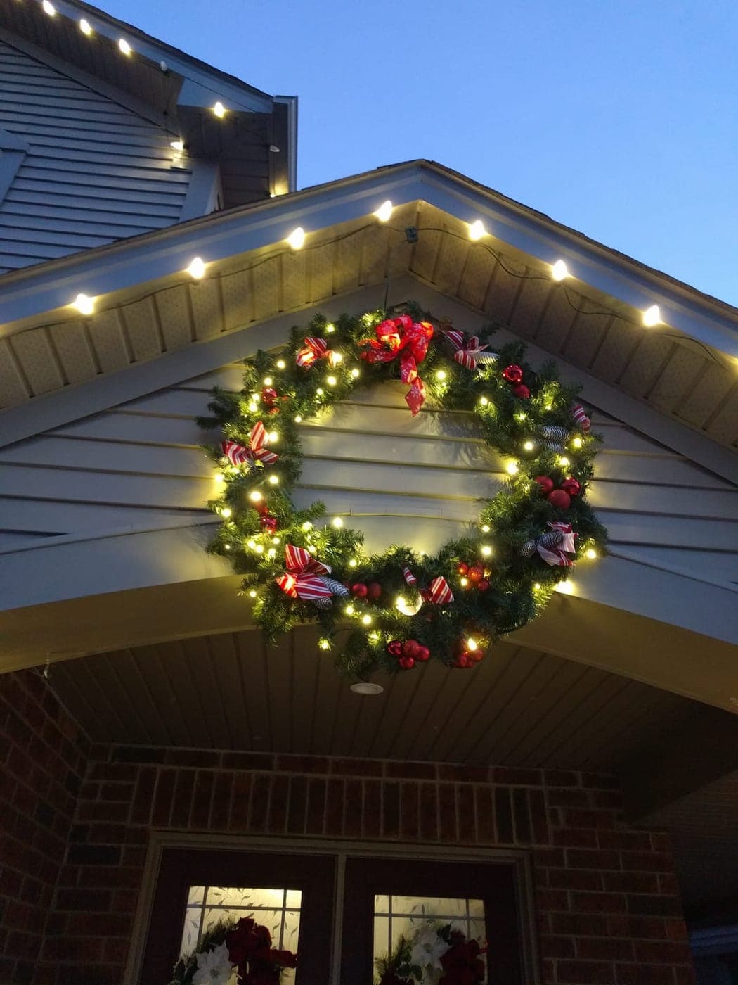 Warm-white lit Christmas wreath with red ribbon accents mounted on a home's gable at dusk.