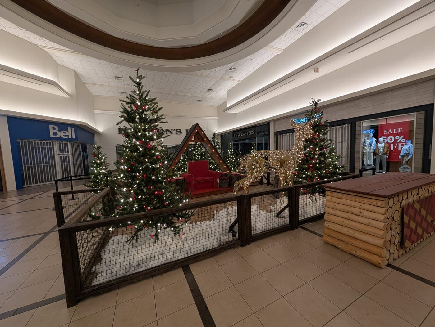 Shopping mall Christmas village display with warm-white lit trees, a glowing reindeer and log-cabin decor