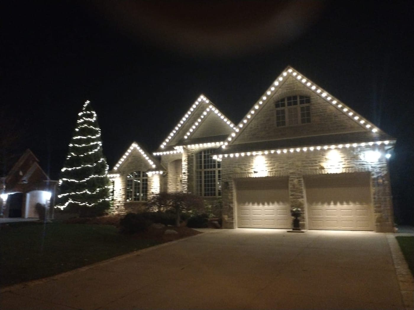 Large stone home outlined in warm-white roofline Christmas lights beside a tall lit spruce tree