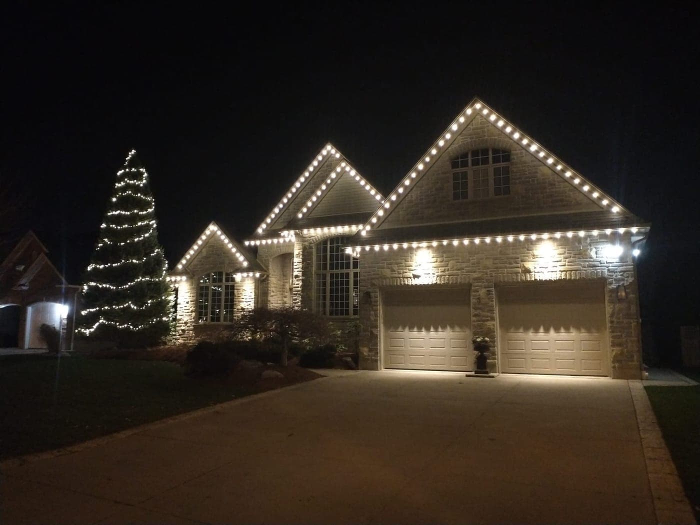 Stone home with warm-white roofline Christmas lights and a tall wrapped evergreen tree at night