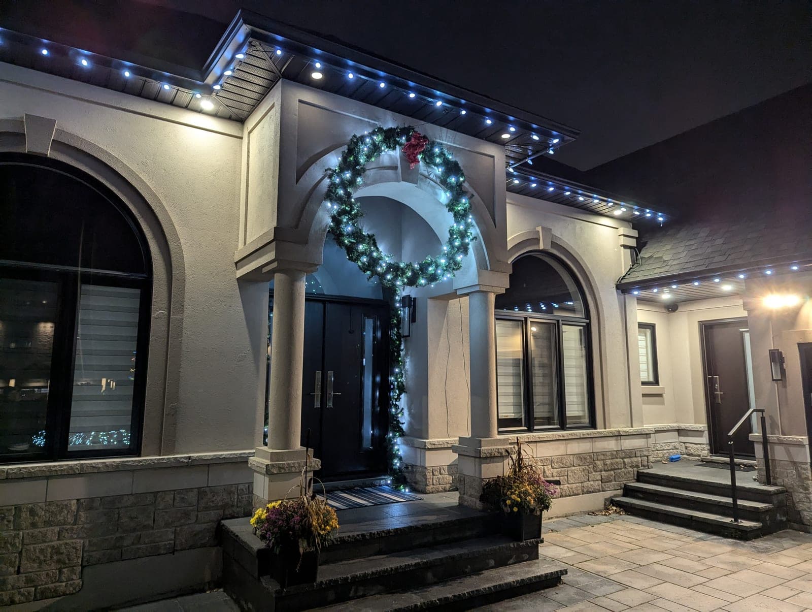 Arched stucco portico decorated with a lit wreath, garland and blue soffit Christmas lights