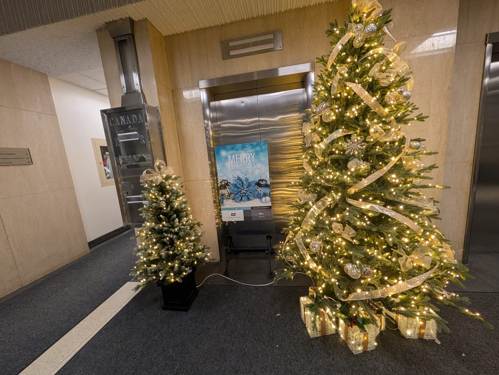 Gold-ribboned commercial lobby Christmas tree with illuminated gift boxes beside the elevators