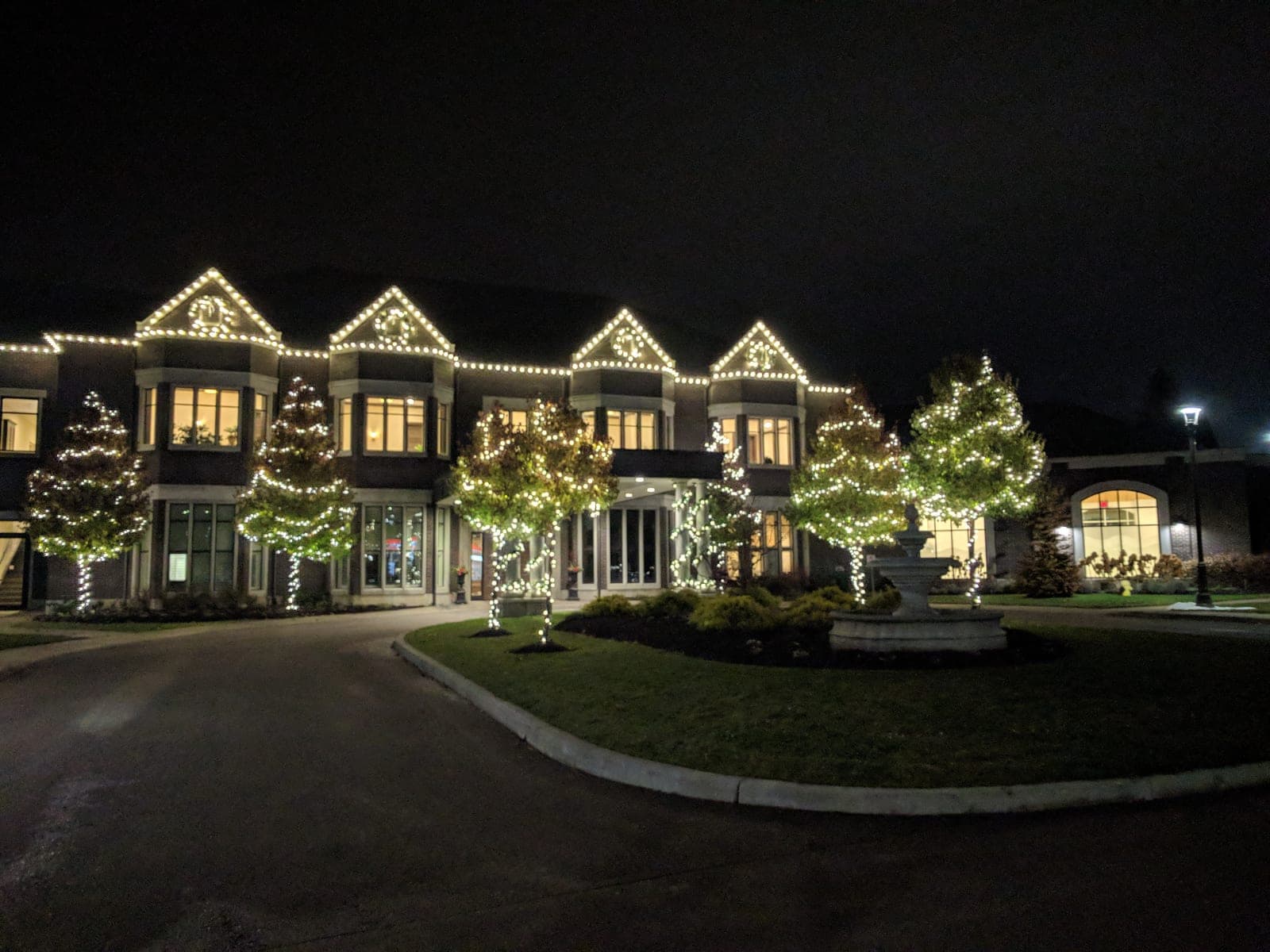 Commercial estate venue with warm-white holiday lighting, a fountain and wrapped trees along the driveway.