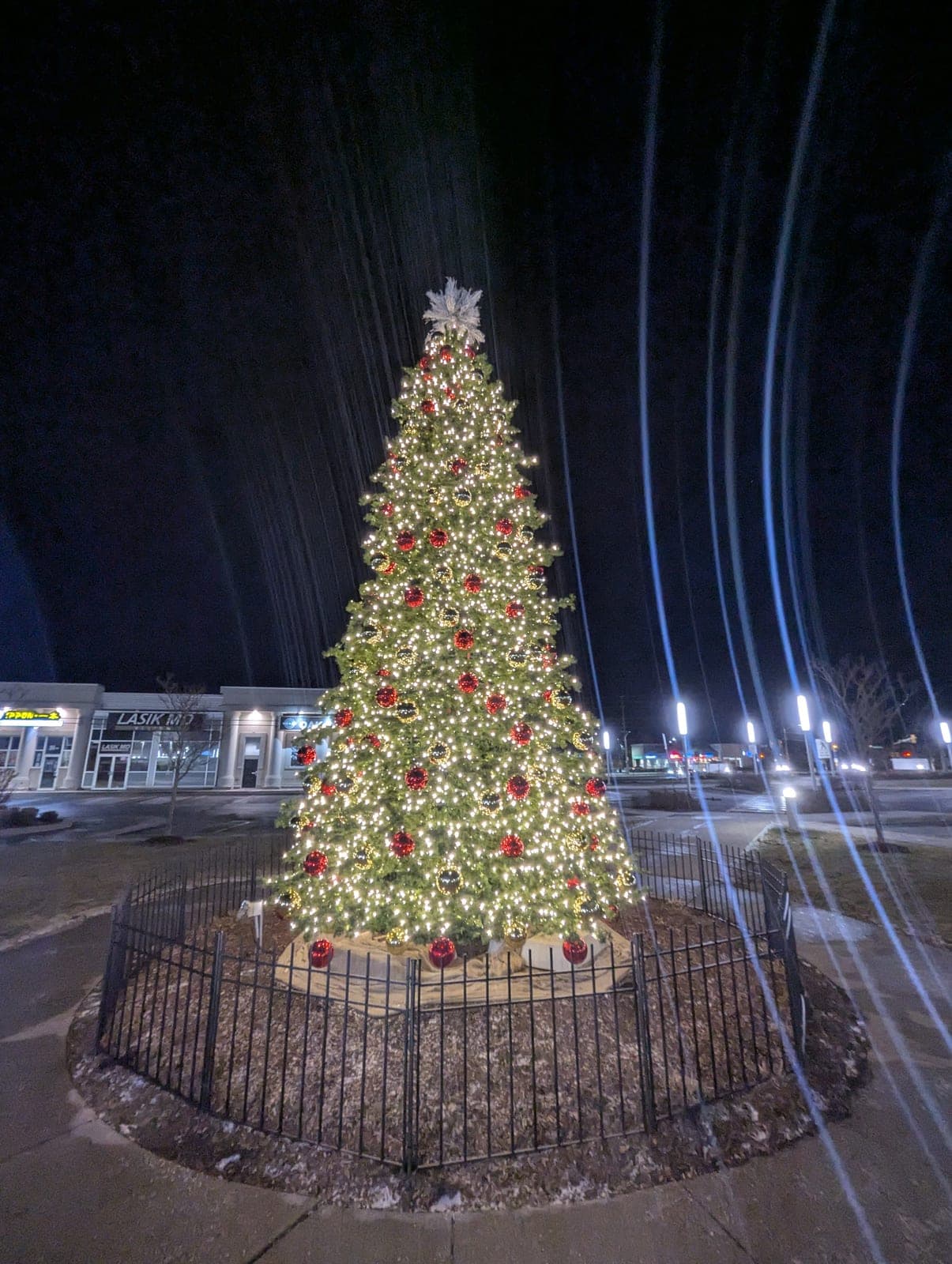 Towering commercial Christmas tree with red and gold ornaments and warm-white lights outside a shopping plaza at night