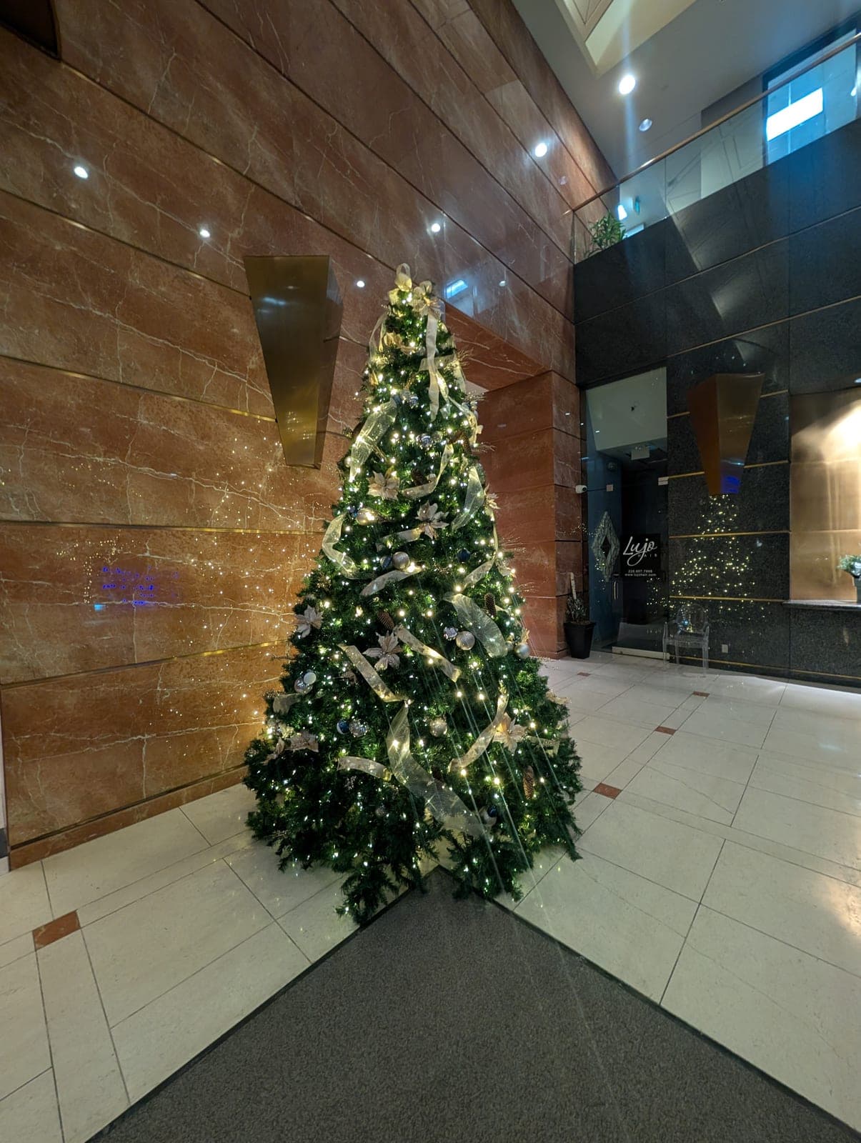 Large Christmas tree with gold ribbon and warm-white lights against a marble feature wall in an office lobby.