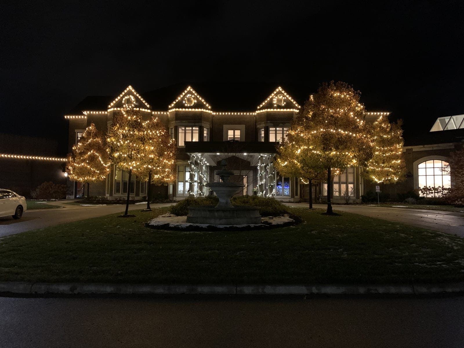 Grand estate lit with warm-white roofline Christmas lights and trees wrapped in lights around an illuminated fountain
