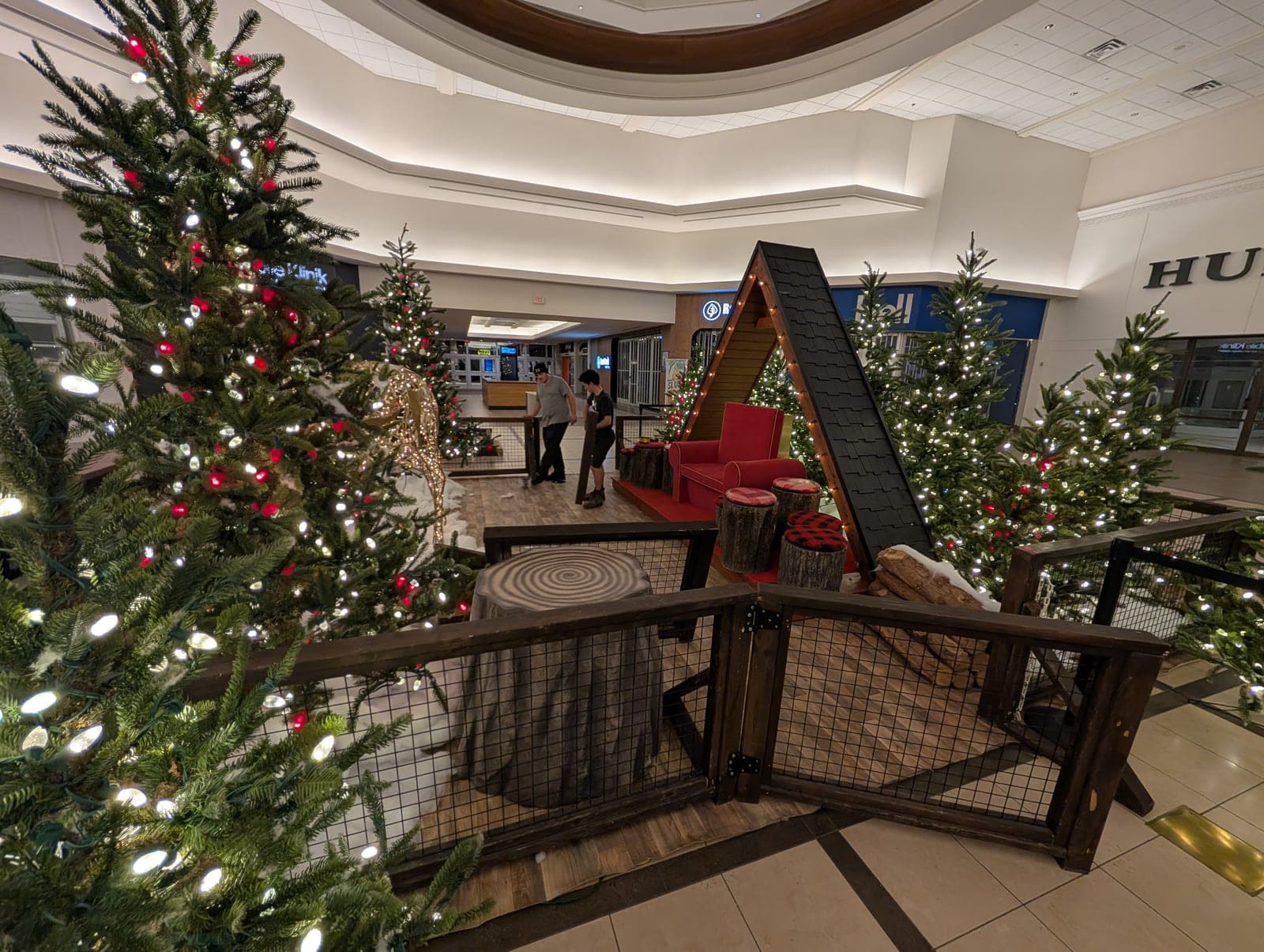 Lit Christmas trees framing a rustic cabin photo set in a mall concourse.