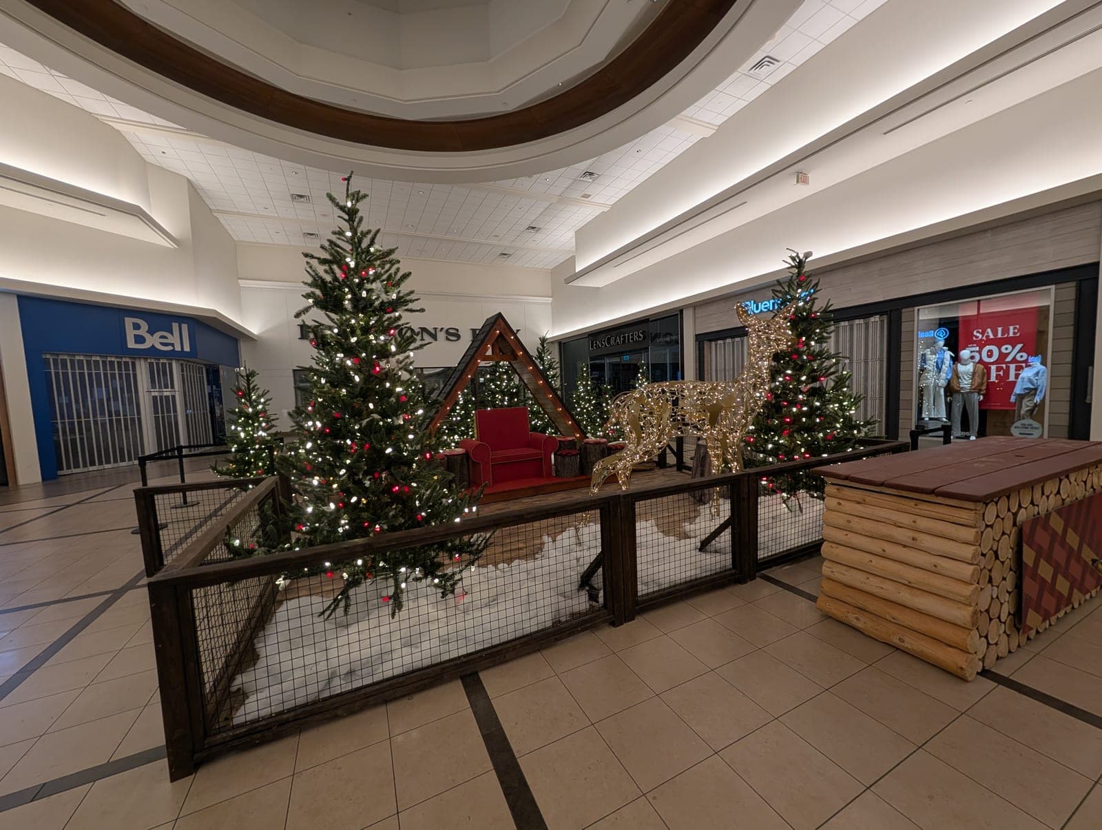 Shopping mall Christmas village display with warm-white lit trees, a glowing reindeer and log-cabin decor