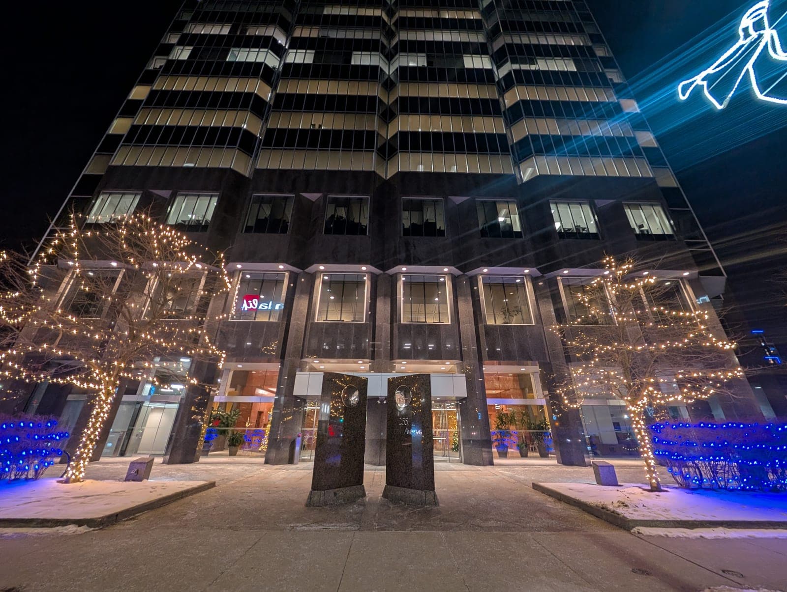 Office tower entrance framed by trees wrapped in warm-white and blue Christmas lights on a winter night.