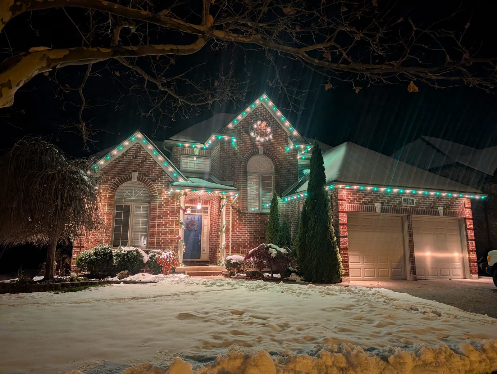 Two-storey brick home with red and green roofline Christmas lights framed by snowy tree branches
