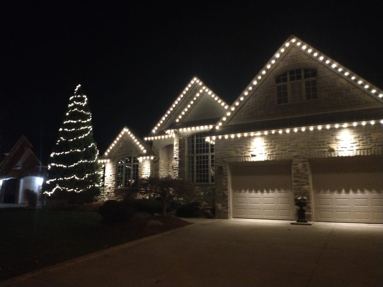 Driveway view of a stone home lined with warm-white roofline Christmas lights and a lit spruce