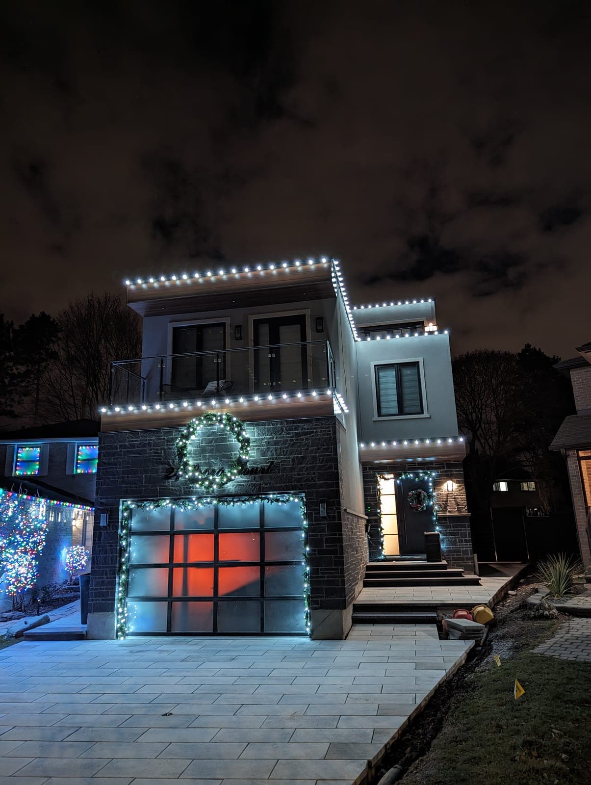 Modern home with warm-white roofline lights, lit wreath and garland-wrapped garage on a snowy street