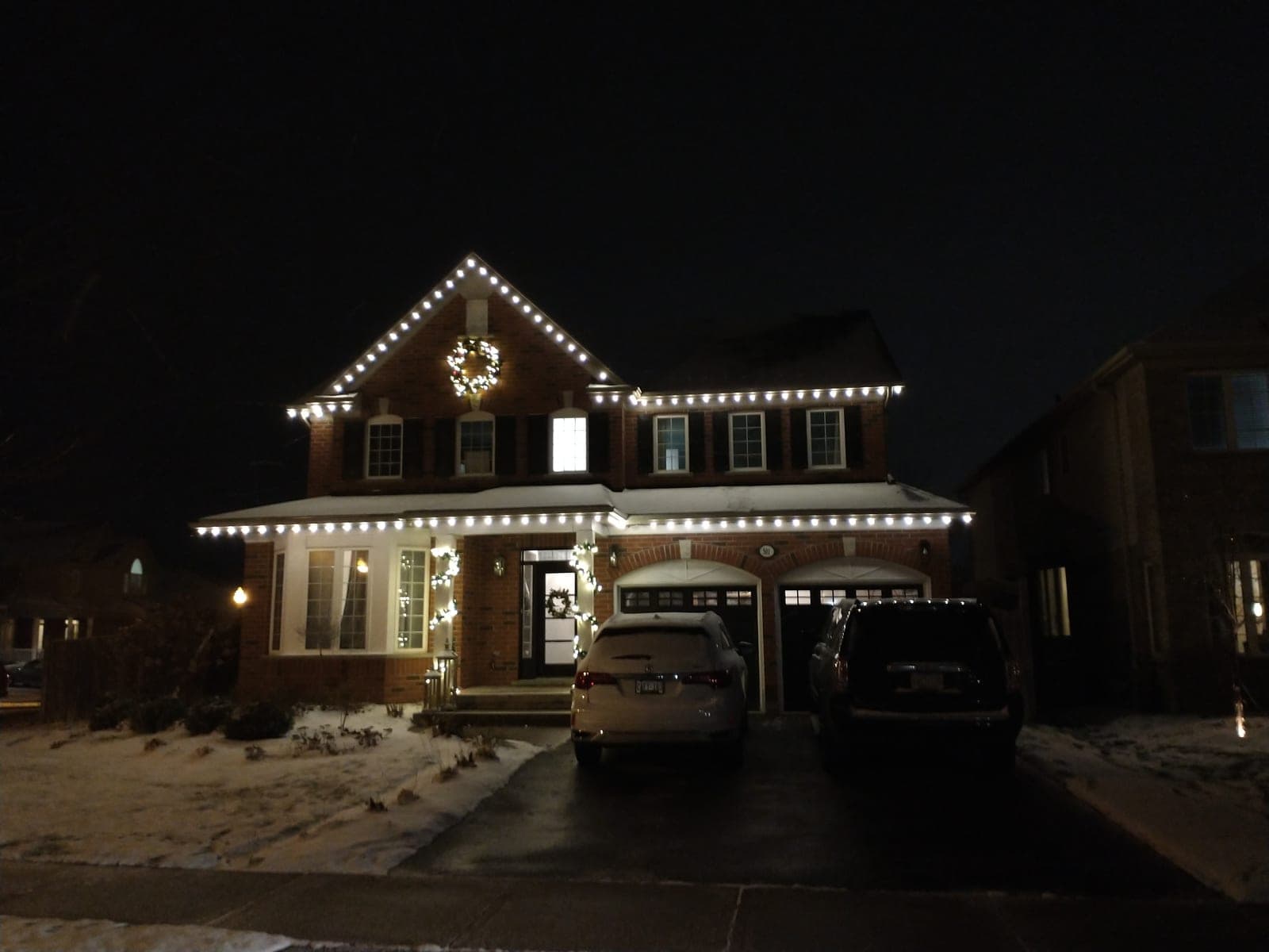 Brick two-storey home with warm-white roofline Christmas lights and a lit wreath on a snowy night