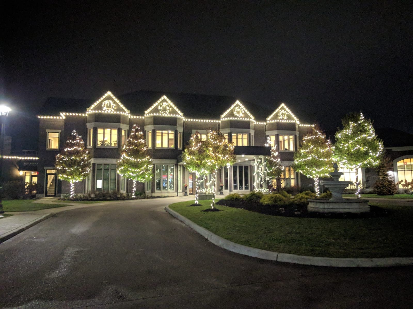 Elegant estate event venue outlined in warm-white roofline lights with lit wrapped trees along the circular drive.